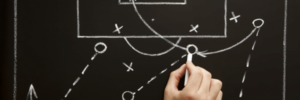 Imagem de um quadro negro com uma mão desenhando a estratégia que pode se utilizar em um jogo de futebol. Essa imagem simboliza o post sobre fazer a estratégia acontecer.