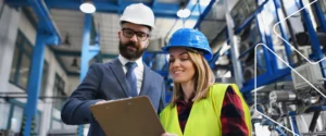 Gestor e técnica com capacetes de segurança revisam uma prancheta em fábrica, representando a aplicação da norma ISO 9000.