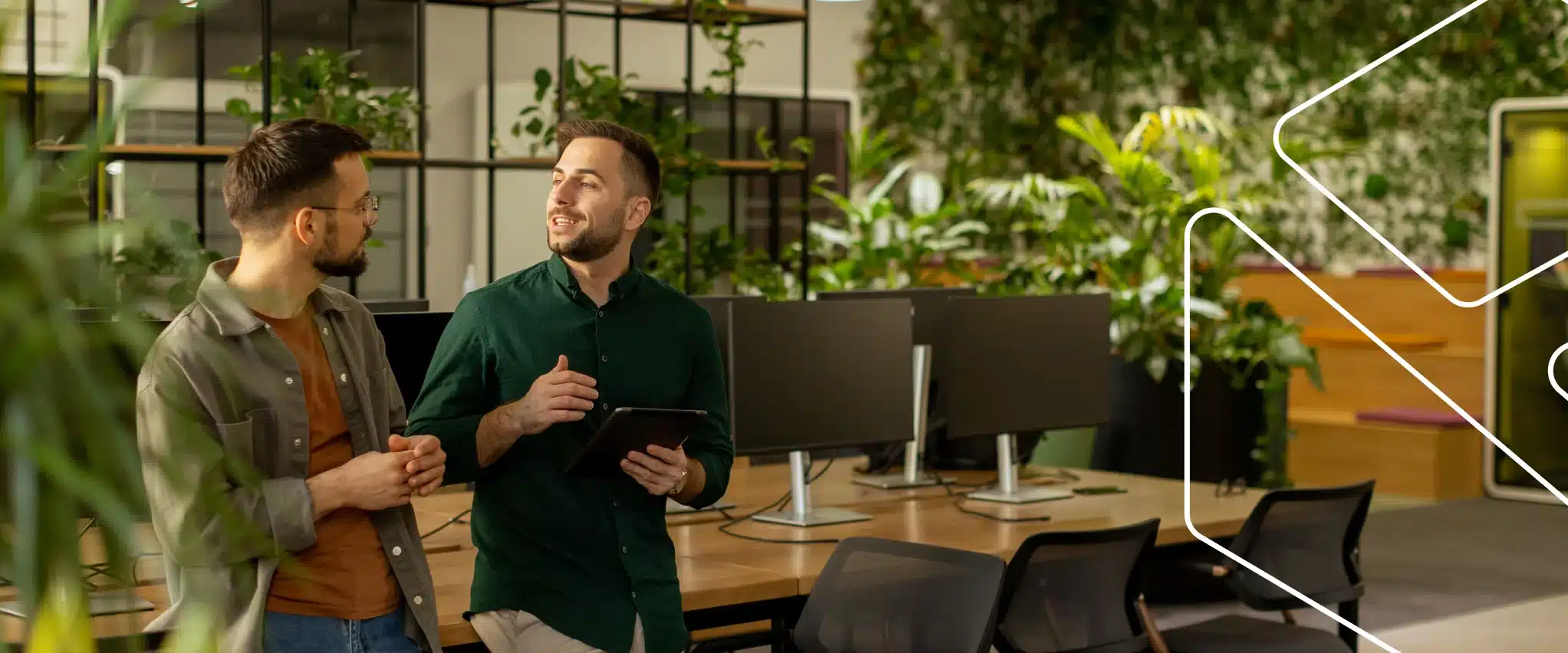 Dois profissionais conversam em um escritório com muitas plantas; um deles segura um tablet