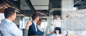 Gestora apresenta um fluxograma no quadro branco para colegas durante reunião de planejamento estratégico em escritório.