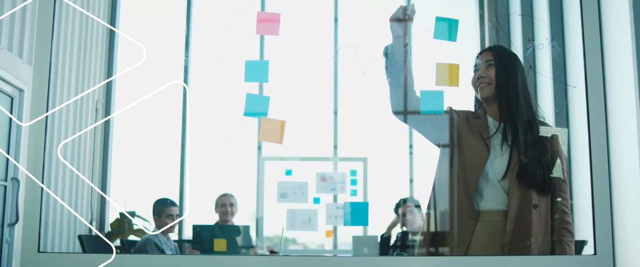Profissional cola post-its em uma parede de vidro durante reunião de planejamento, observada por três colegas sentados à mesa.
